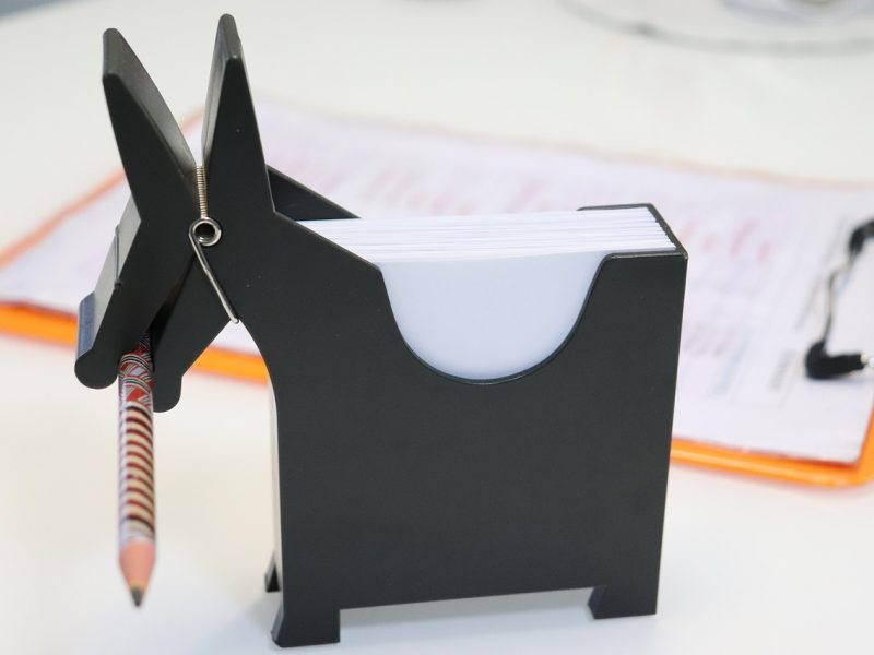 https //kanju3.ai/ 编辑在米色办公桌前用铅笔在纸质清单上逐条圈画待核名称，旁边贴着黄色与珊瑚色便签，桌面有放大镜与记号笔